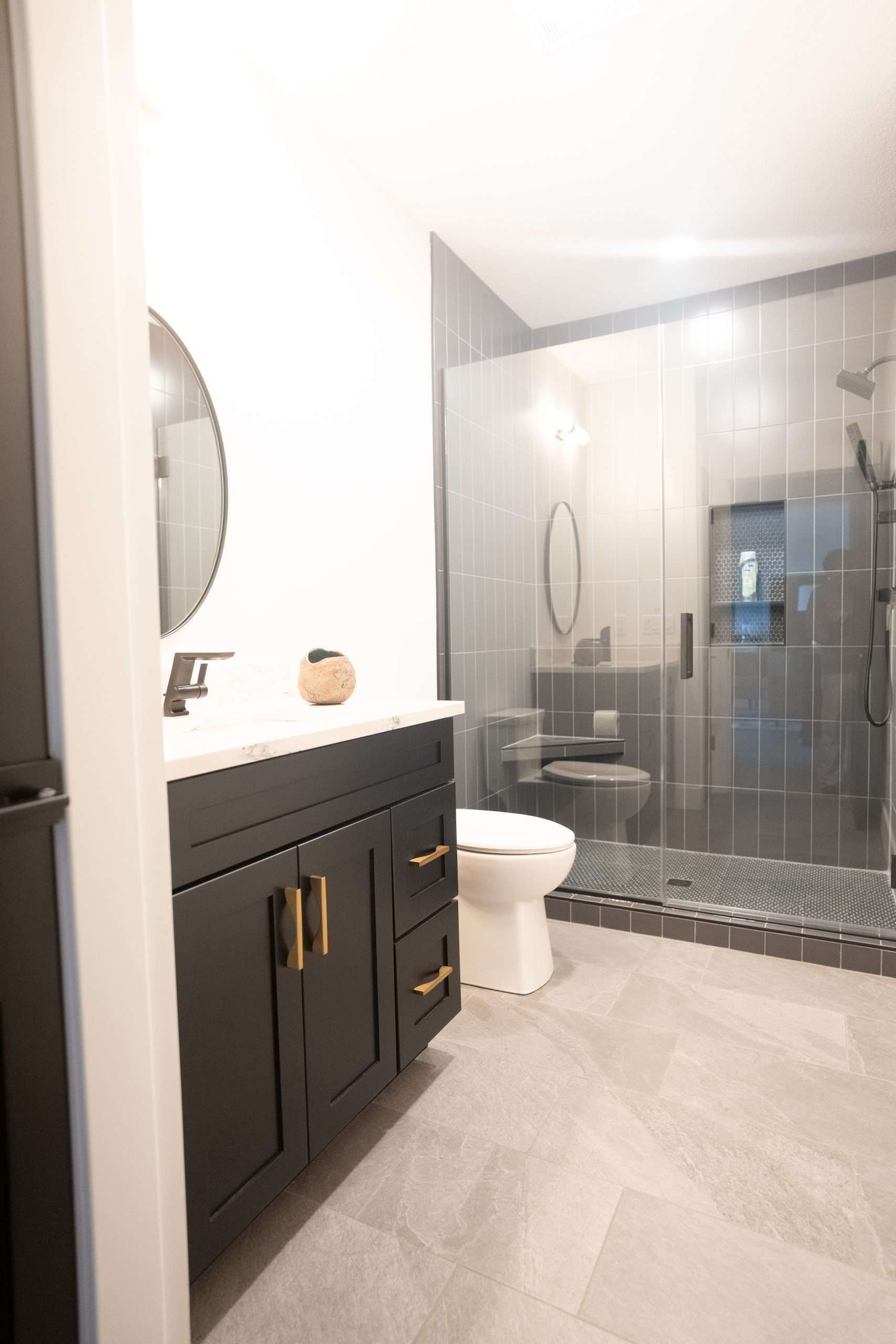 Modern bathroom with dark vanity, white toilet, and glass shower enclosure with black tile.