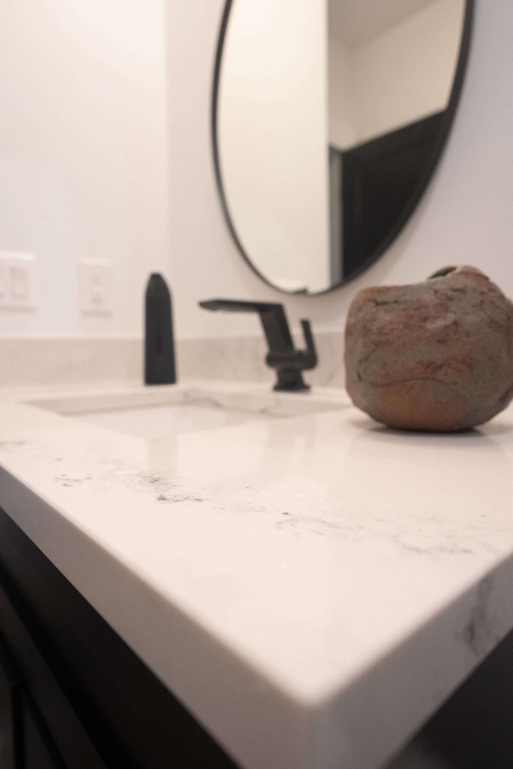 Bathroom vanity with a round mirror, black faucet, soap dispenser, and a stone countertop decor piece.