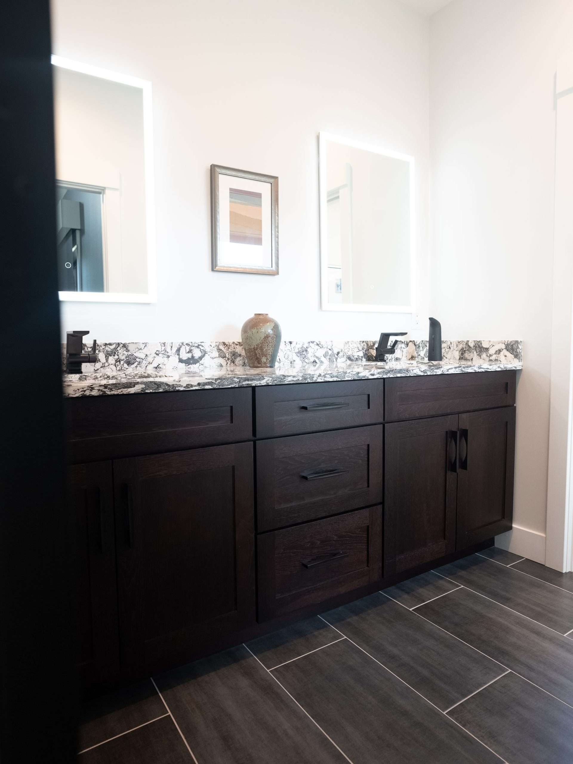Dark wood bathroom vanity with granite countertop, mirror, and patterned floor tiles in a bright white room