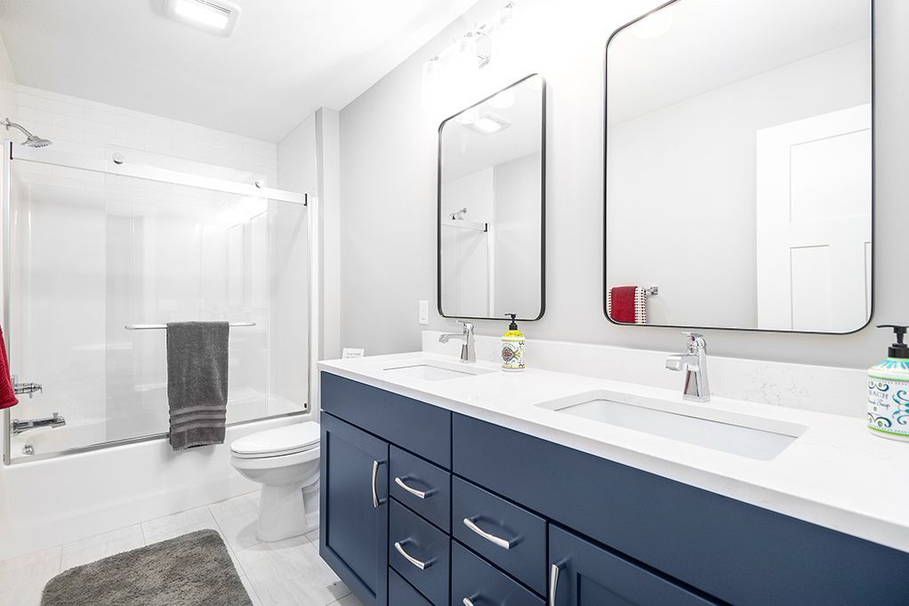 Bright white bathroom with blue vanity, double mirrors, sink, toilet, and shower enclosure.