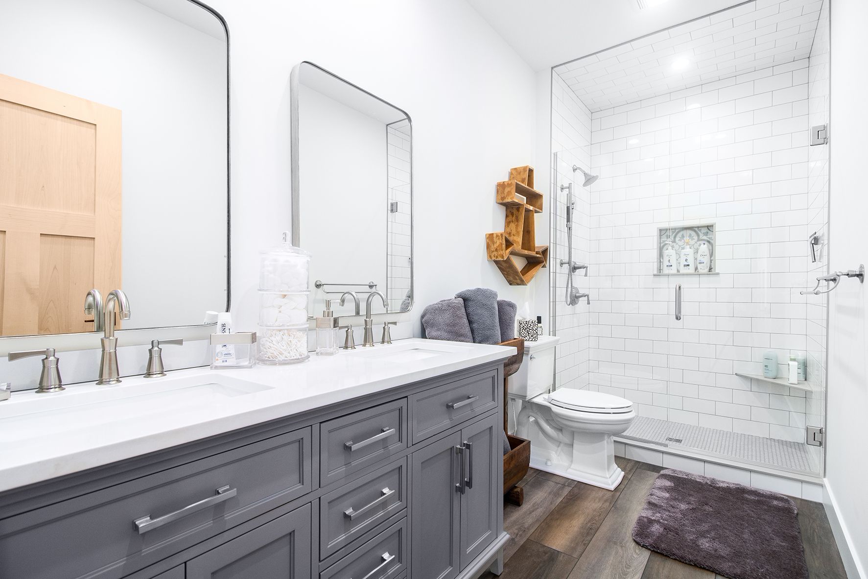 Bright modern bathroom with gray vanity, large mirror, toilet, glass shower, and dark rug