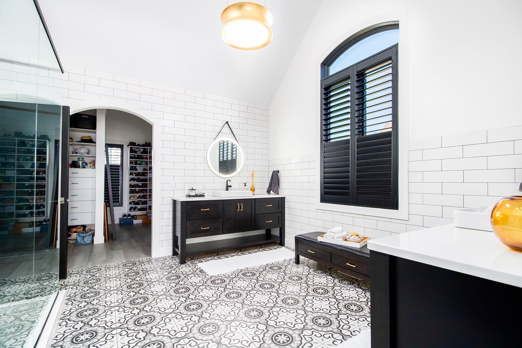 Bright white bathroom with black accents, patterned tile floor, vanity, arched mirror, and window shutters
