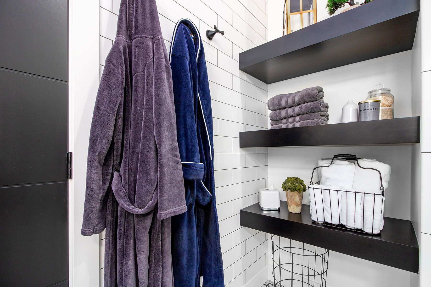 Bathroom with two robes hanging on a wall, shelves holding towels and toiletries, and a small plant.