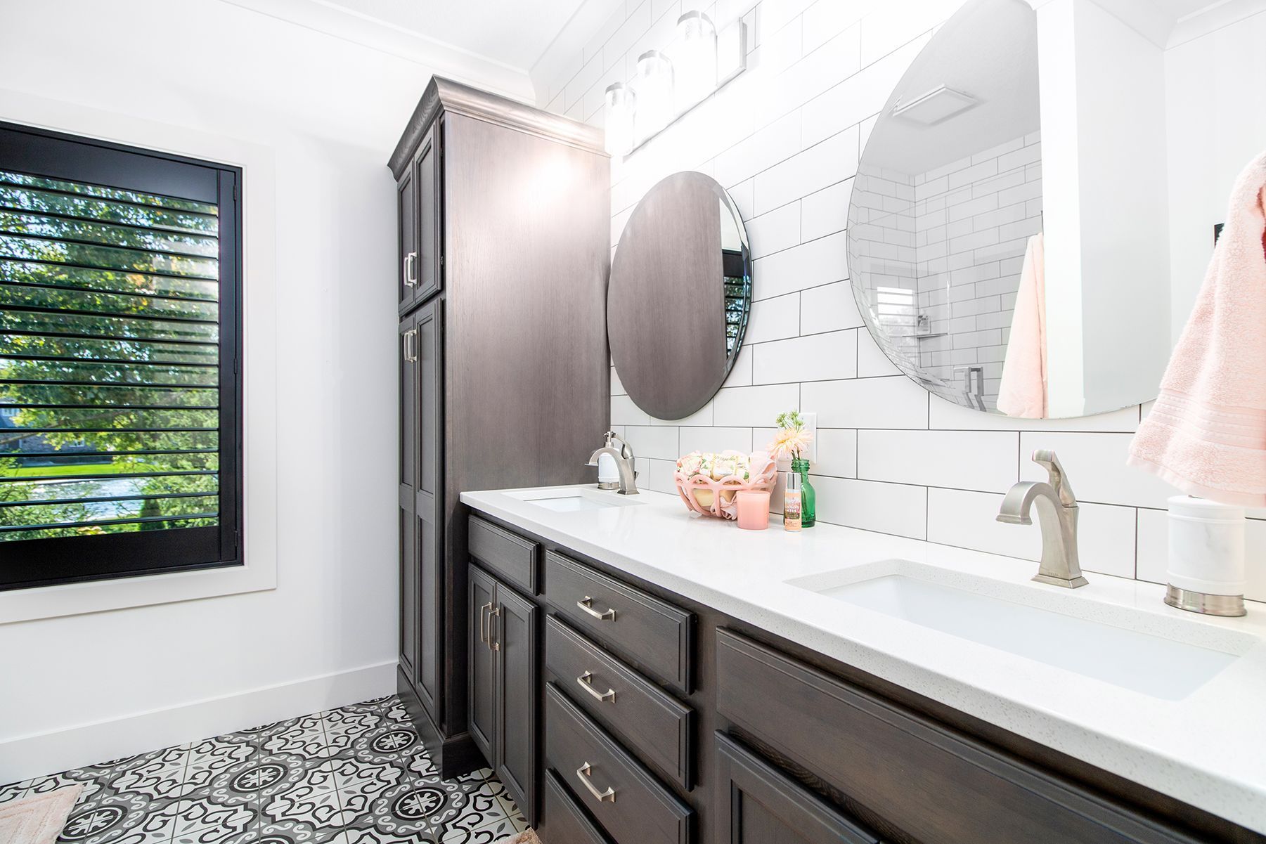 Bright modern bathroom with dark wood vanity, oval mirrors, and a window with greenery outside