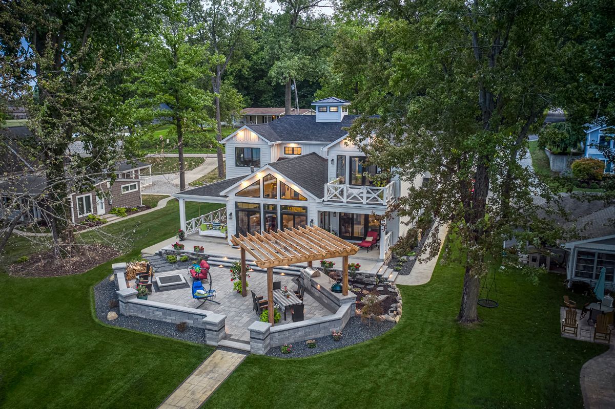 Aerial view of a large stone house with a terraced backyard patio, pergola, and lush trees.