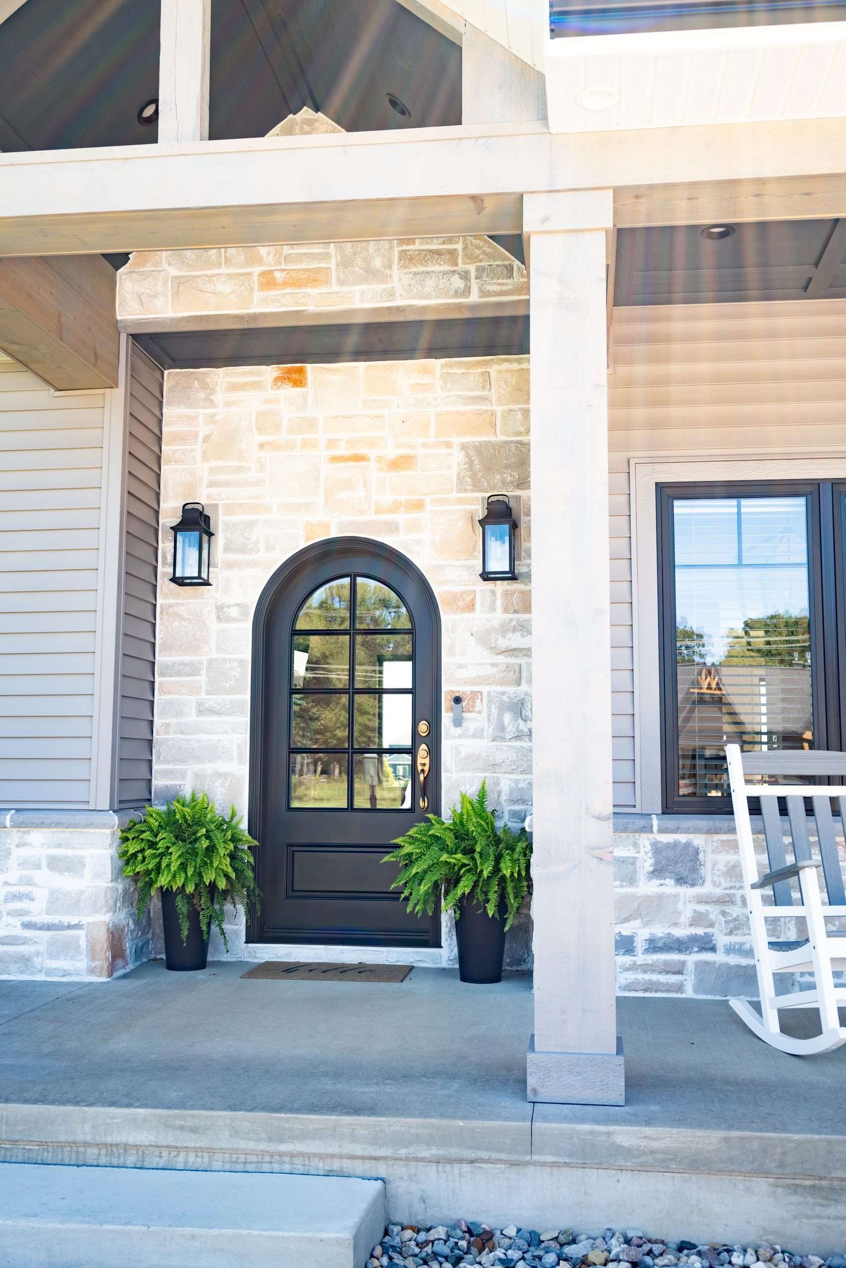 Front porch with a black arched door, white siding, potted plants, and a rocking chair