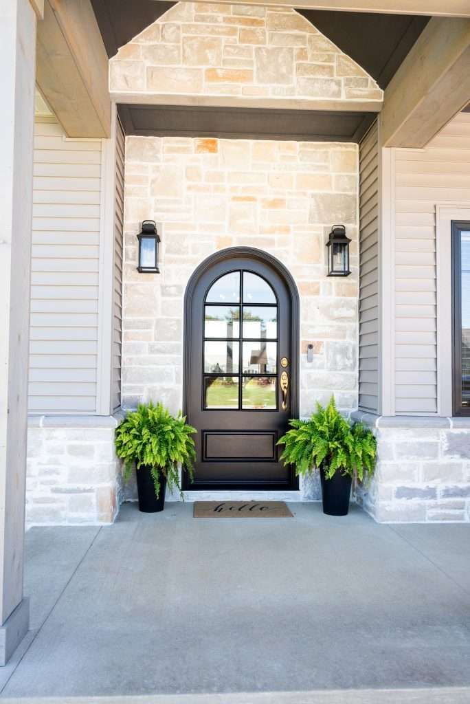 Front entry with arched black door, lanterns, potted plants, and light stone siding