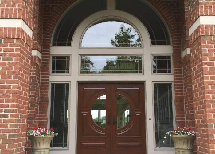 Brick house front entrance with arched window and double wooden doors, flanked by potted flowers.