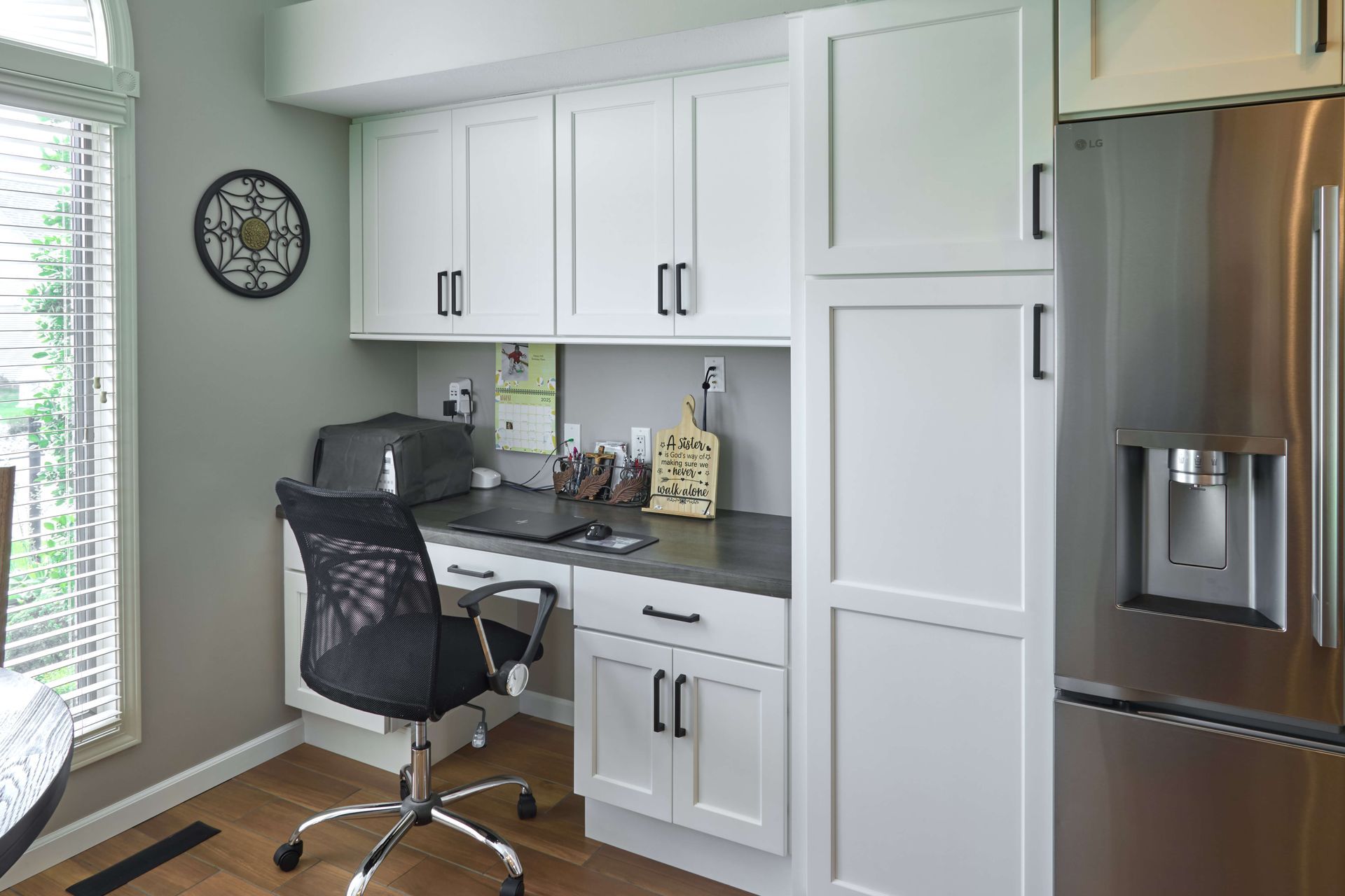 Home office nook with white cabinets, desk, black chair, and stainless steel refrigerator.