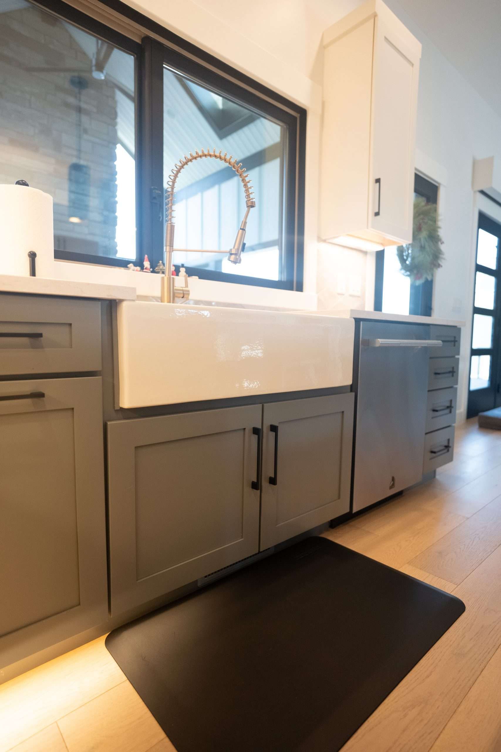 Modern kitchen sink with gray cabinets, white backsplash, black rug, and sunlight by a window