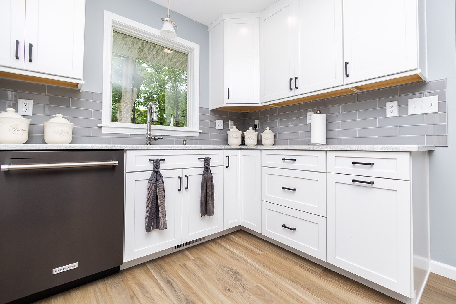 Bright white kitchen with black dishwasher, gray tile backsplash, window, and wood floor