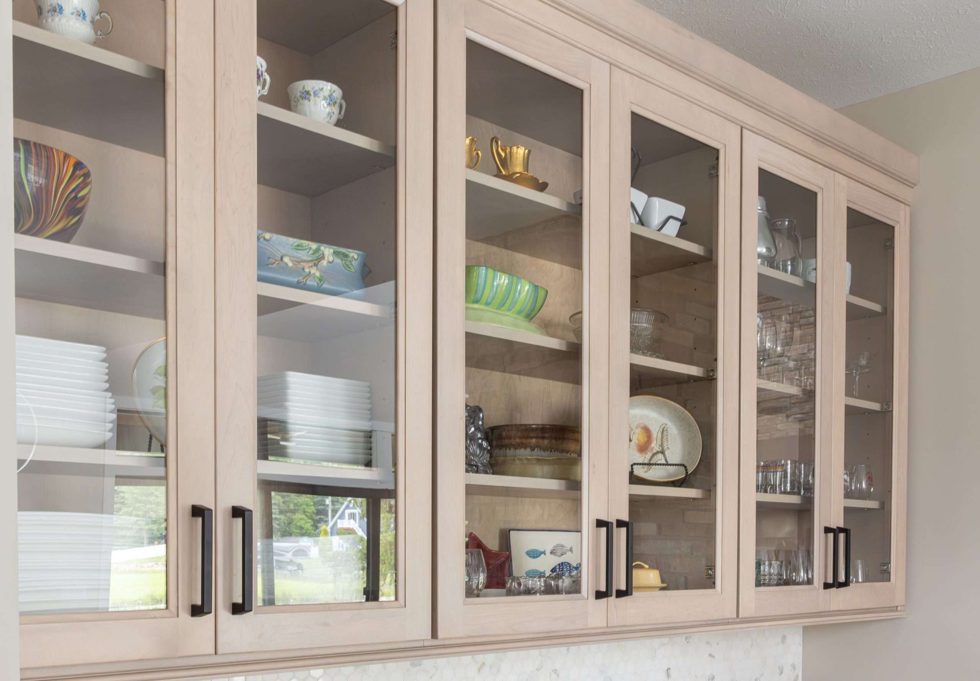 Glass-front kitchen cabinets displaying dishes, bowls, and glassware in a beige kitchen.