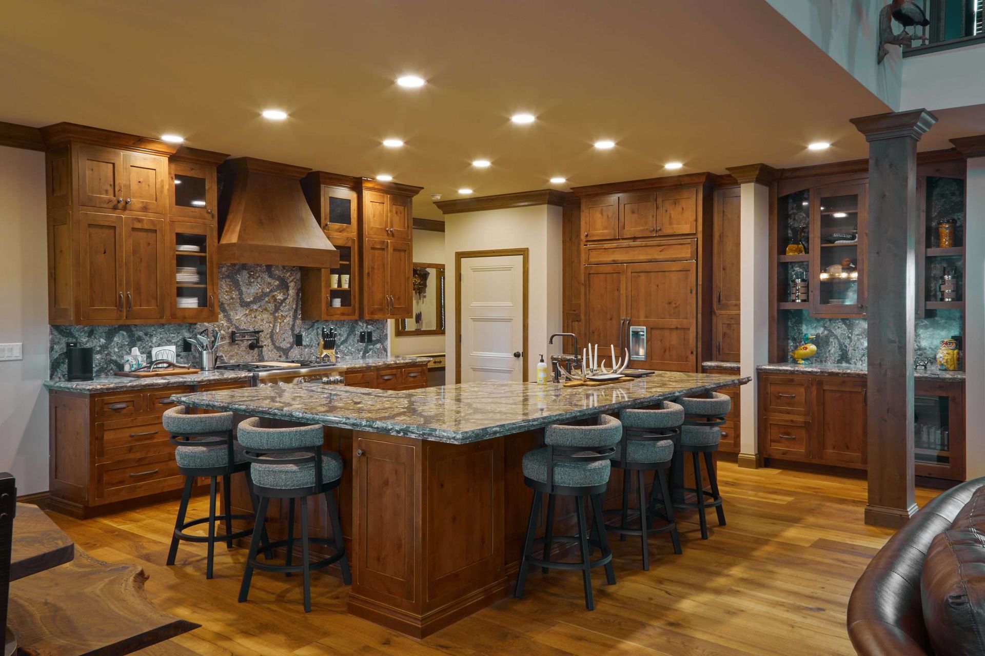 Warm kitchen with granite island, wooden cabinets, bar stools, and recessed ceiling lights.