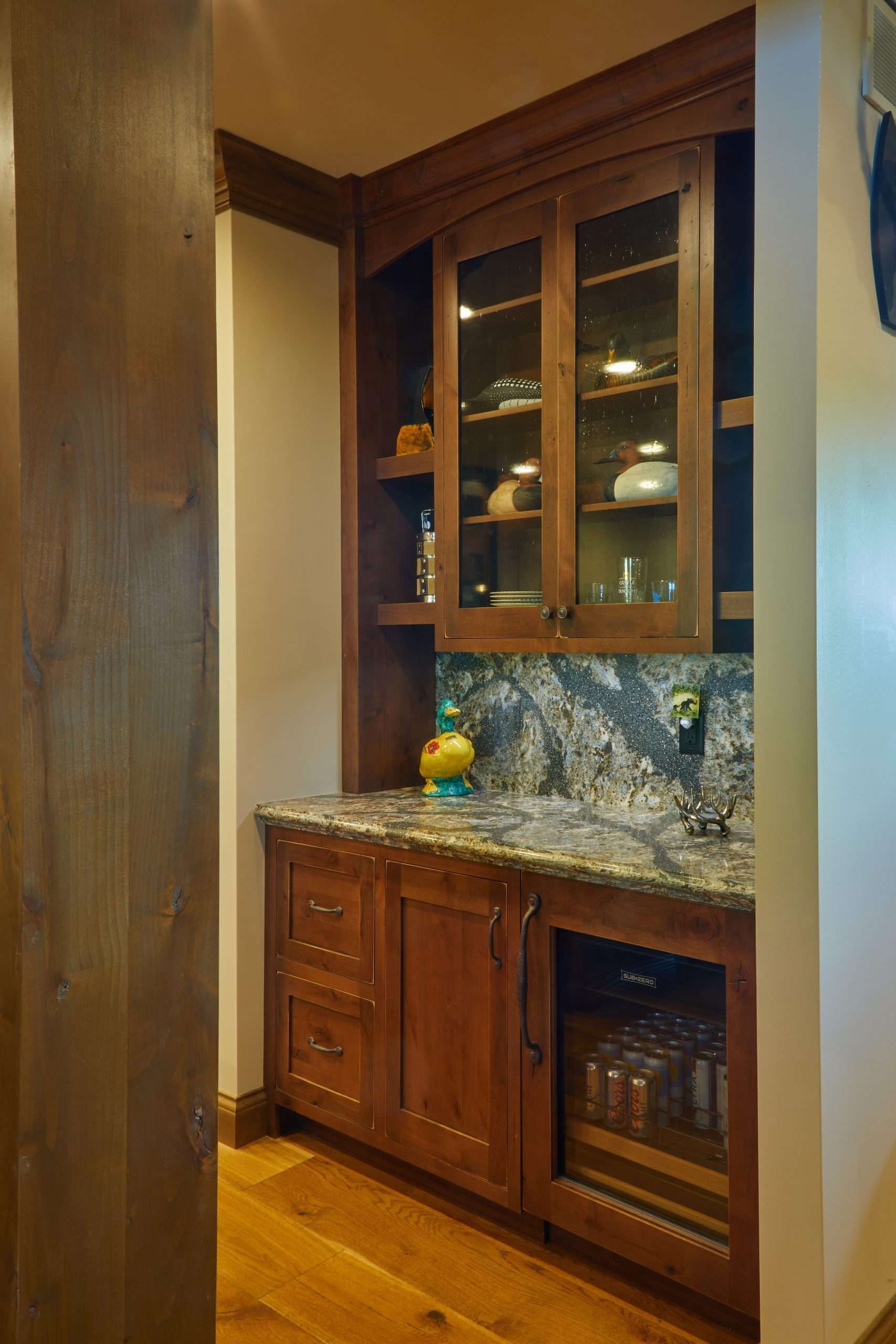 Wooden kitchen nook with granite countertop, glass-front cabinets, and wine cooler under warm lighting