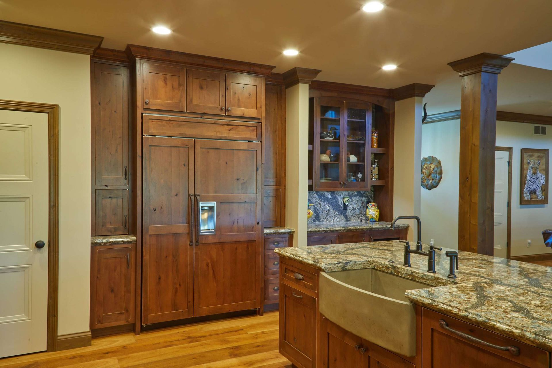 Warm kitchen with wood cabinets, granite island, stainless sink, and built-in refrigerator under recessed lights