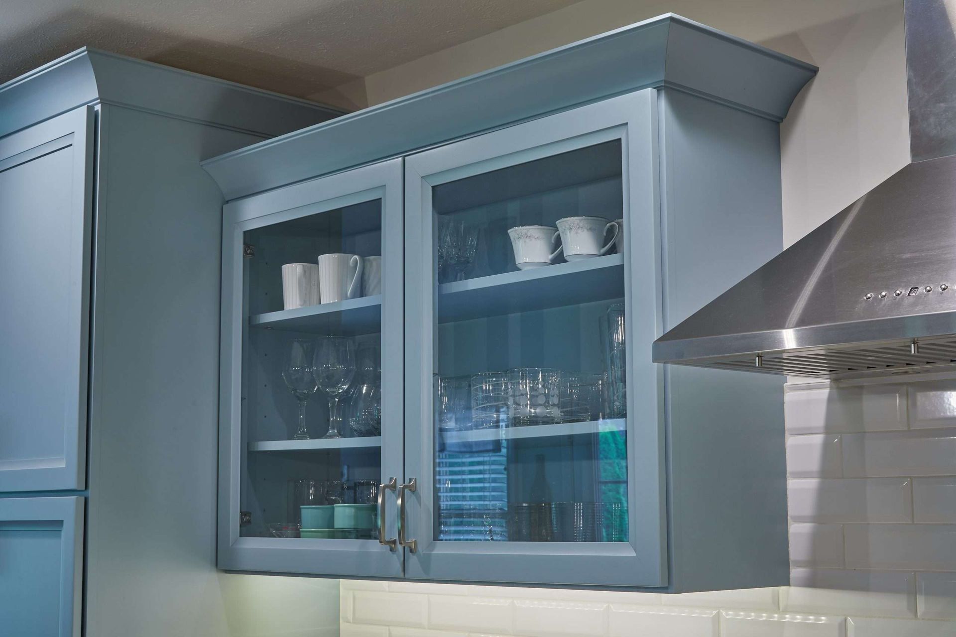 Blue kitchen cabinets with glass doors displaying dishes and glasses next to a stainless steel range hood.