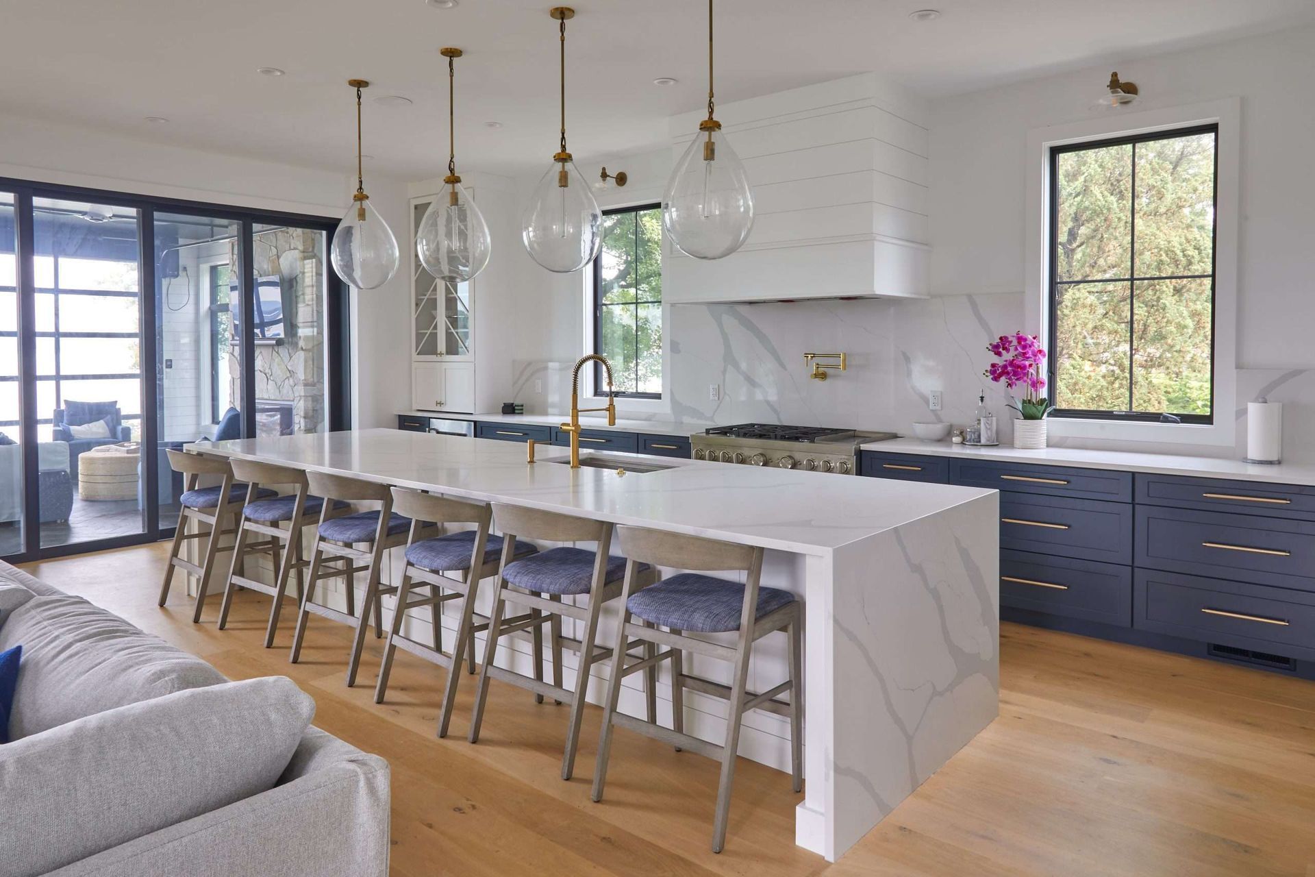 Modern kitchen with large marble island, pendant lights, bar stools, and blue cabinetry under bright windows