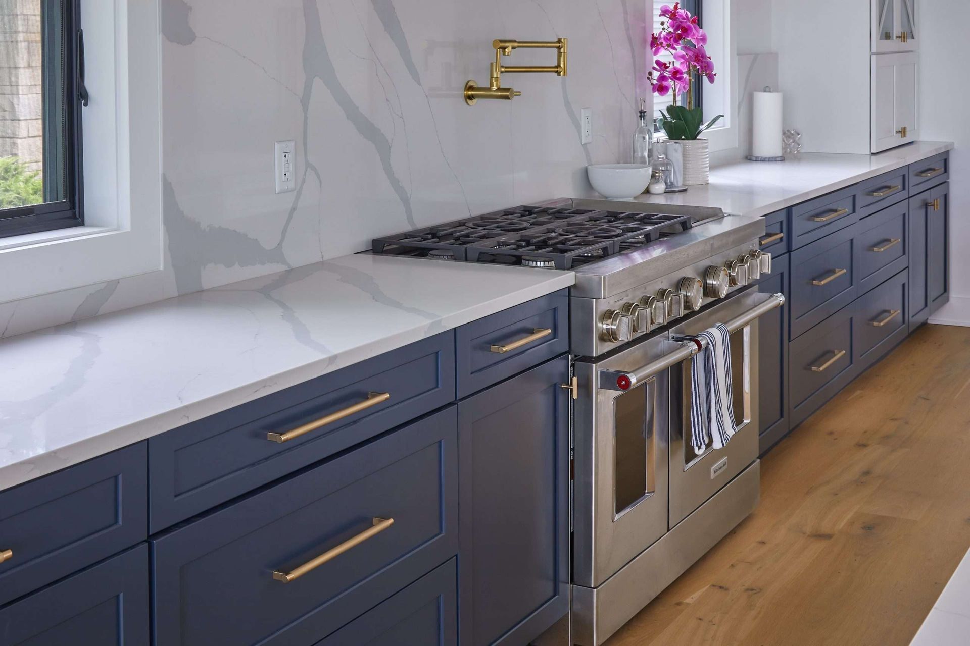 Modern kitchen with navy cabinets, white countertops, stainless stove, and gold faucet along a bright window.