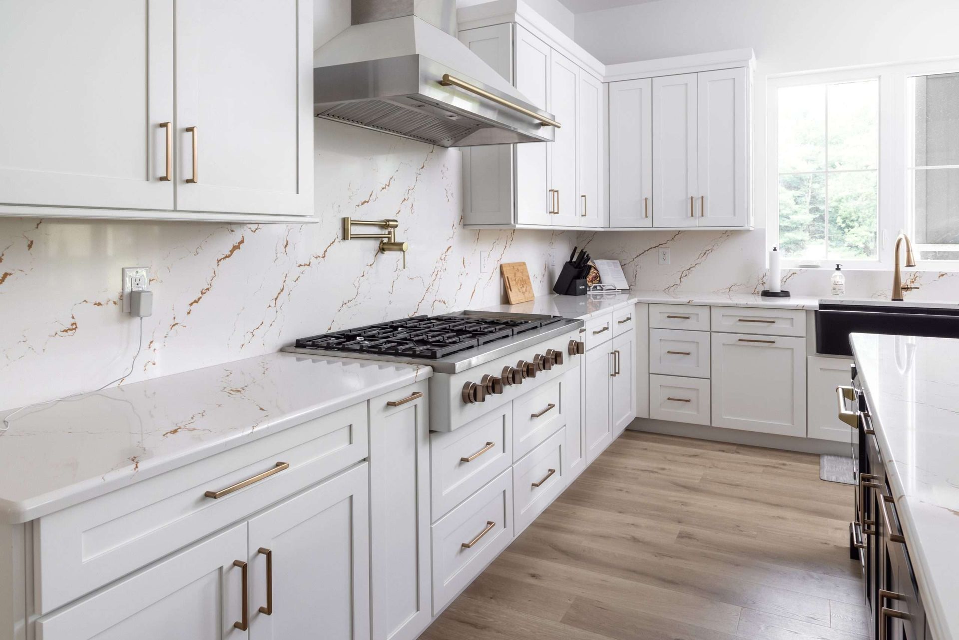 Bright modern white kitchen with gold handles, marble counters, wood floor, and stainless steel stove