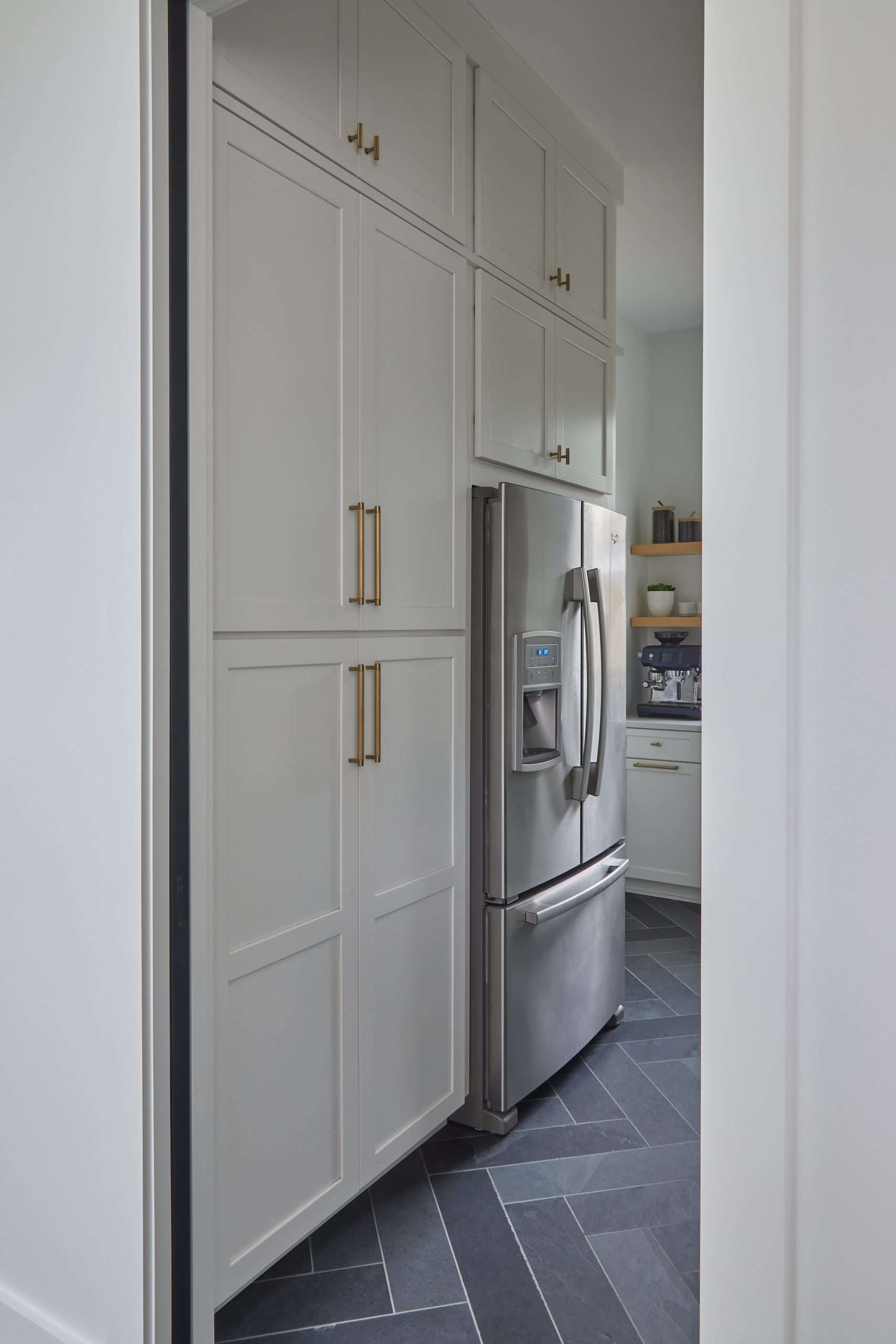 Narrow kitchen with tall white cabinets, stainless steel refrigerator, and dark patterned floor.