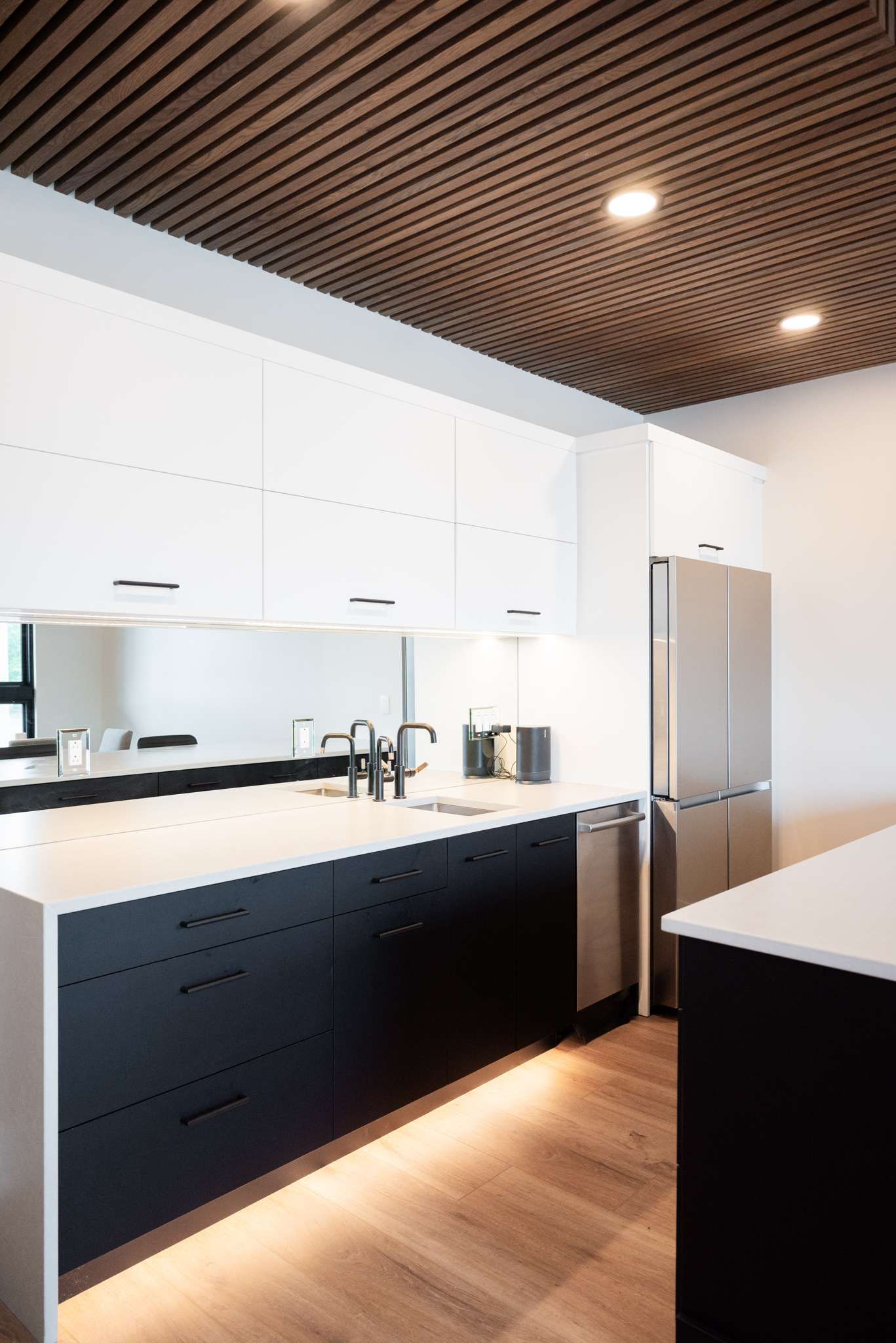 Modern kitchen with dark wood ceiling, white cabinets, black lower drawers, and stainless steel fridge.