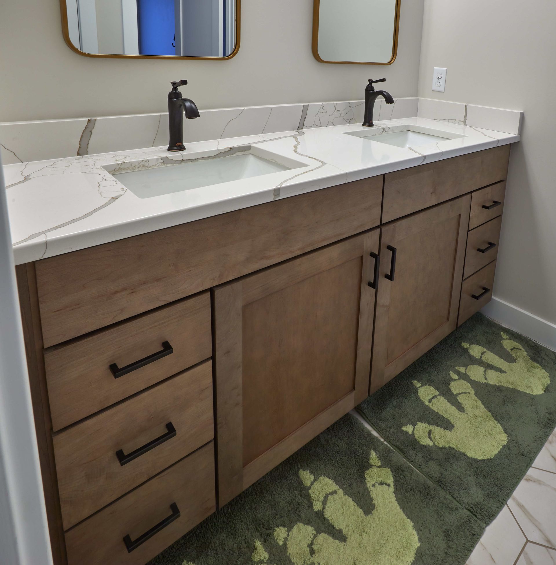 Double vanity bathroom sink with wood cabinets, black faucets, and green patterned rug