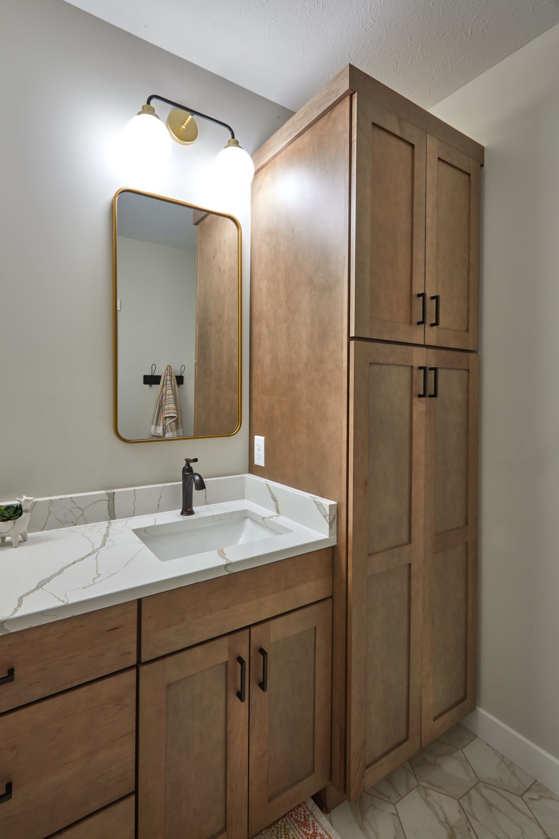 Small bathroom vanity with white countertop, wood cabinets, mirror, light fixture, and tall storage cabinet.