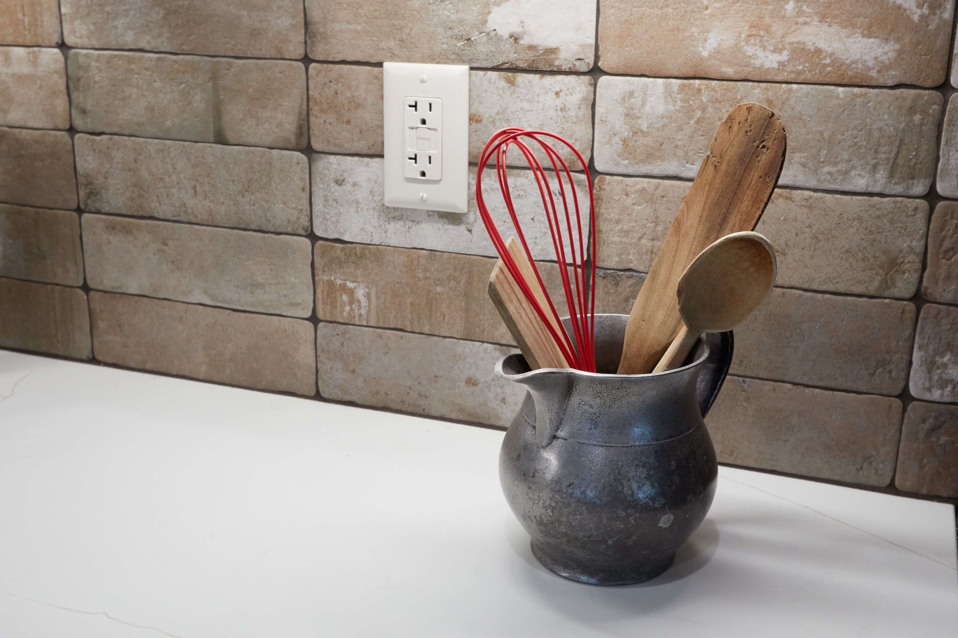 Kitchen countertop with a gray utensil holder, red whisks, wooden spoons, and a tiled backsplash.