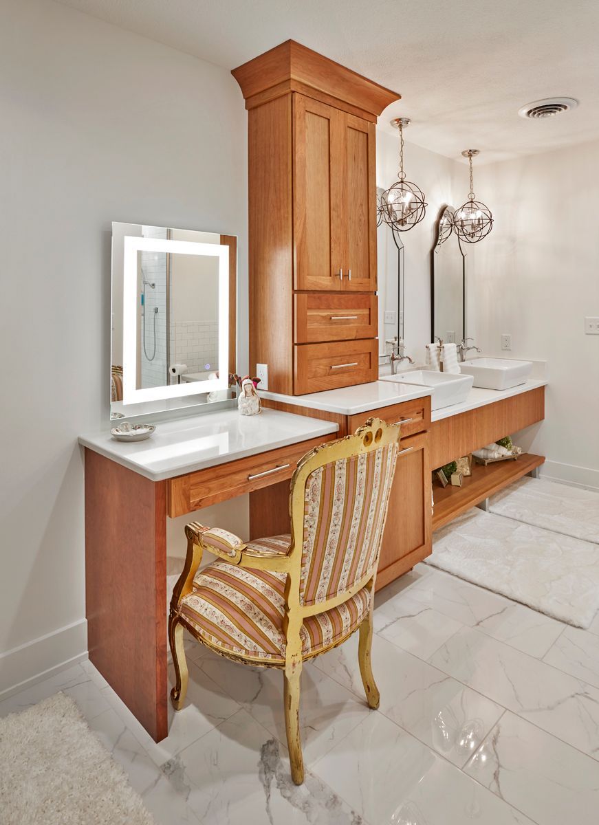 Luxury bathroom vanity with wood cabinets, white marble counters, mirrors, and a gold chair