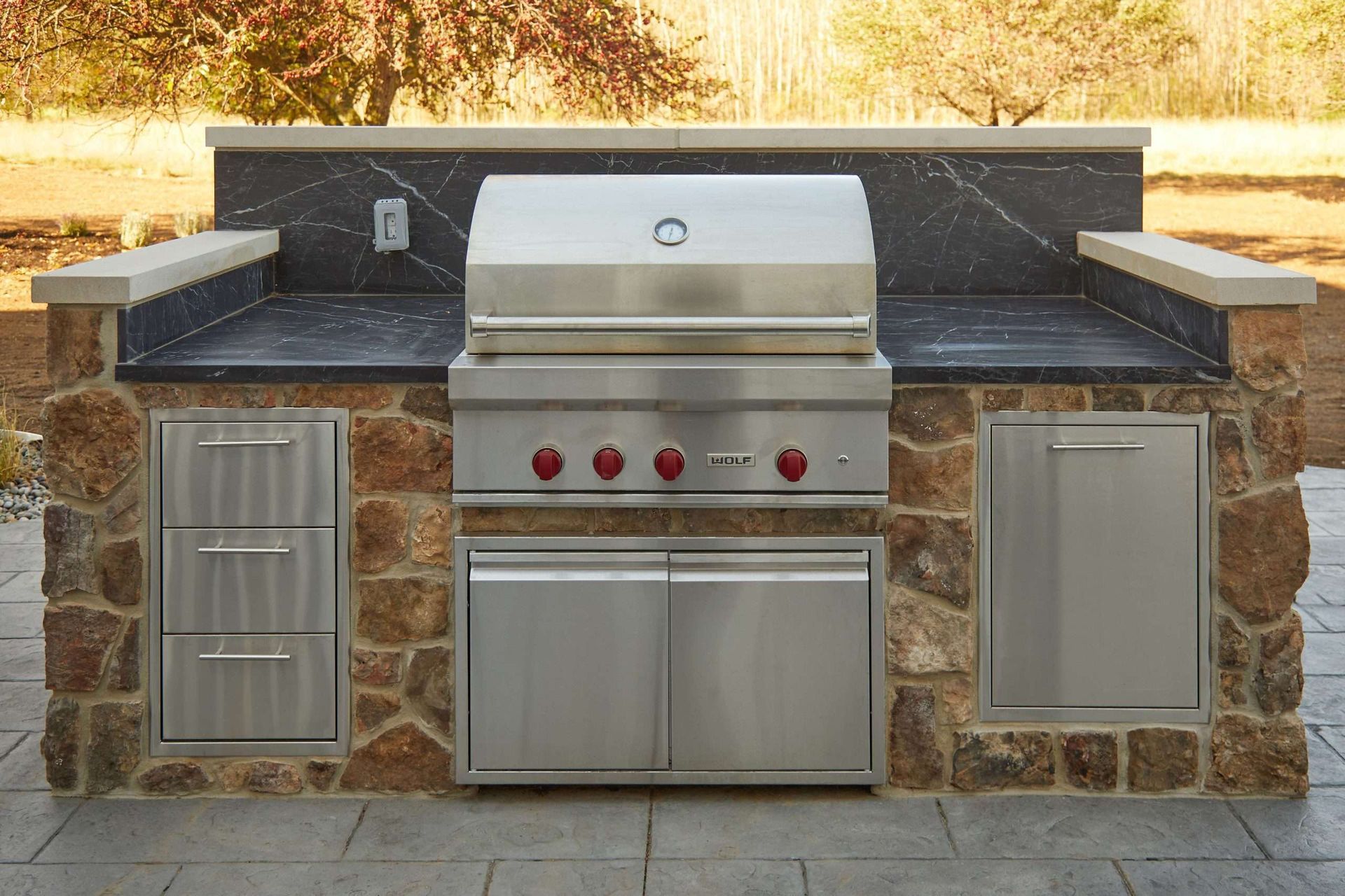 Outdoor stone barbecue grill with stainless steel doors, red knobs, and side countertops in a backyard