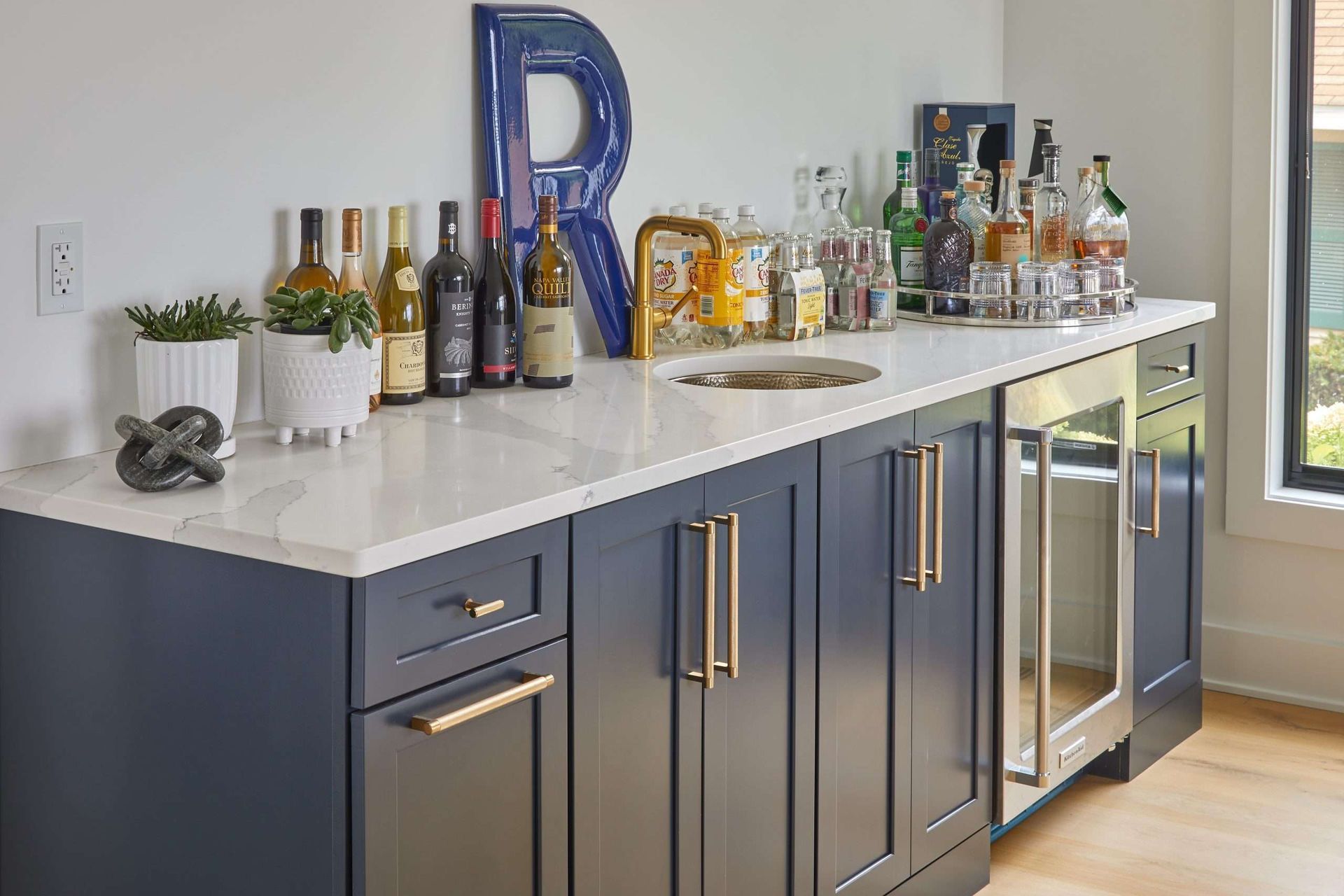 Modern bar area with dark cabinets, white countertop, bottles, glasses, and a large blue letter R.