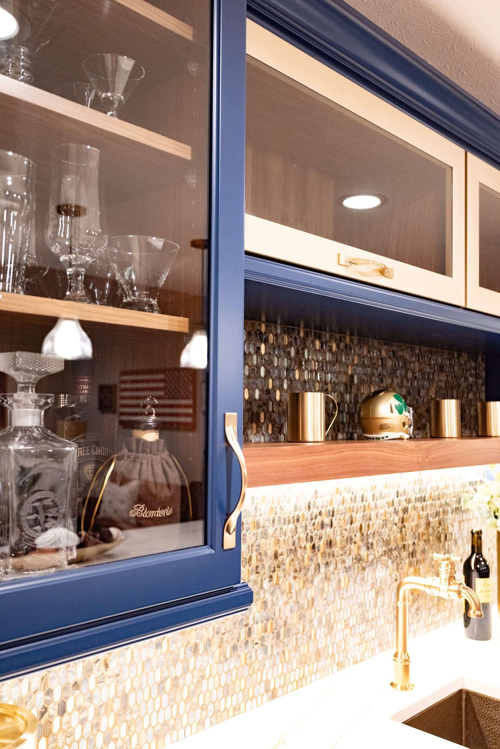 Blue kitchen cabinets with glassware and shelves above a sink and gold faucet, lit under a backsplash light