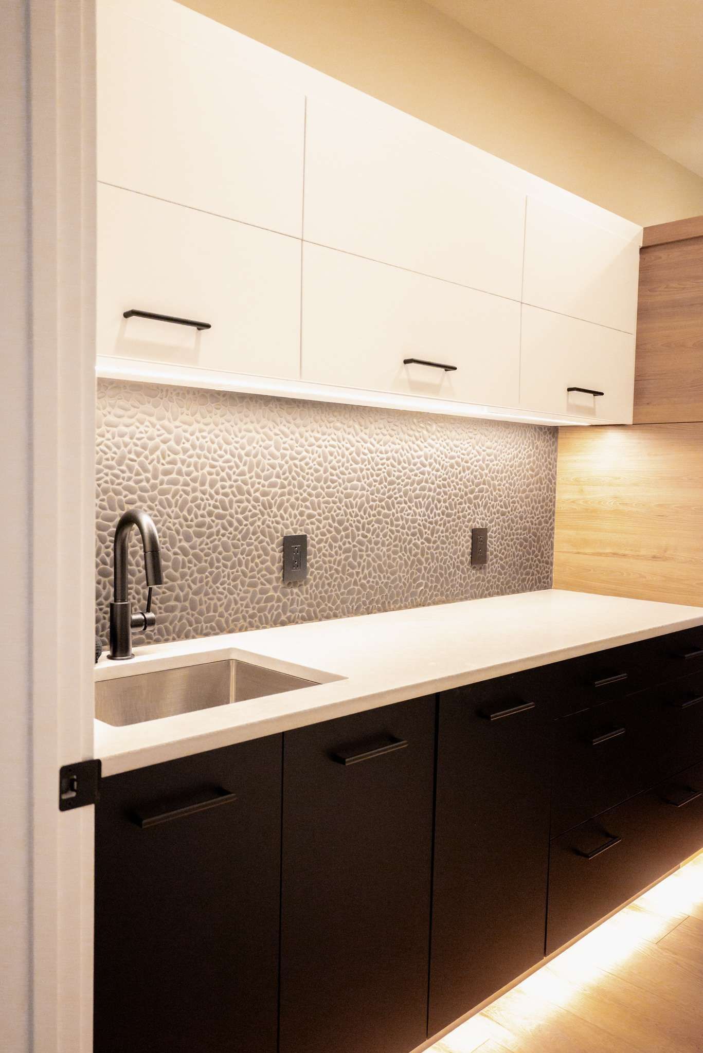 Modern kitchen with white upper cabinets, dark lower cabinets, sink, mosaic backsplash, and under-cabinet lighting.