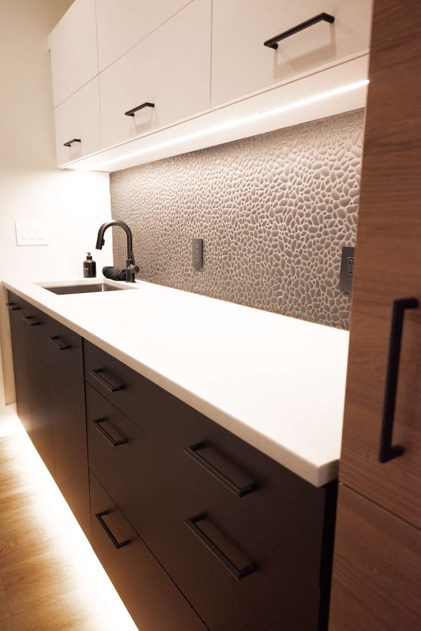 Modern kitchen with white countertop, dark cabinets, under-cabinet lighting, and tiled backsplash.