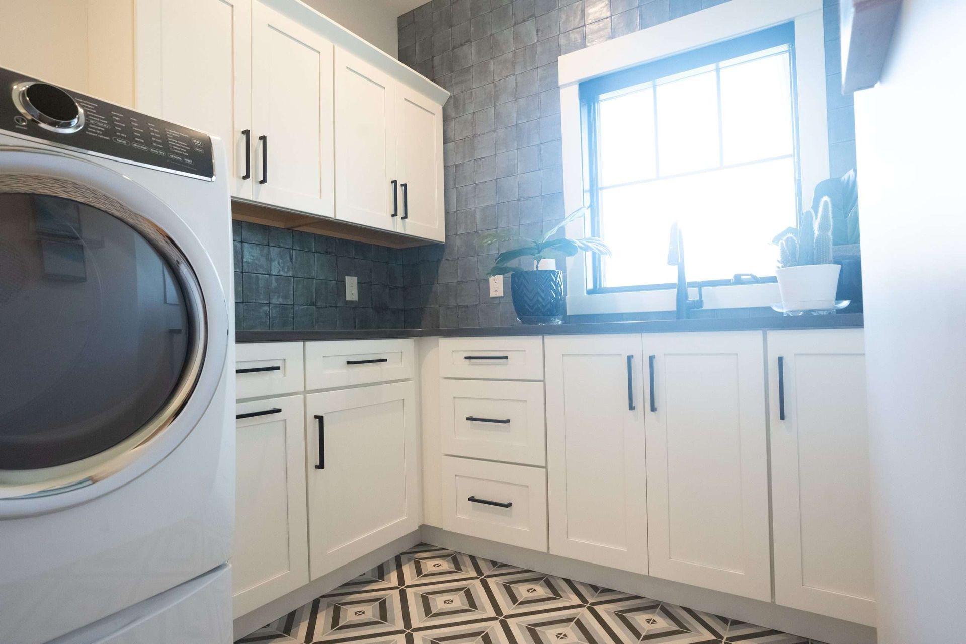 Bright laundry room with white cabinets, a front-loading washer, black tile backsplash, and a sunlit window above the sink.