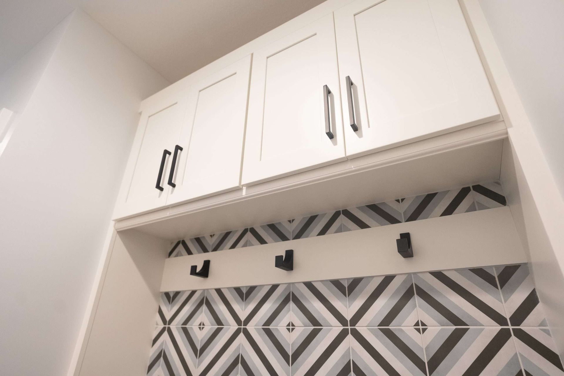 White wall cabinets above a black-and-white geometric tile backsplash and coat hooks in a narrow entryway