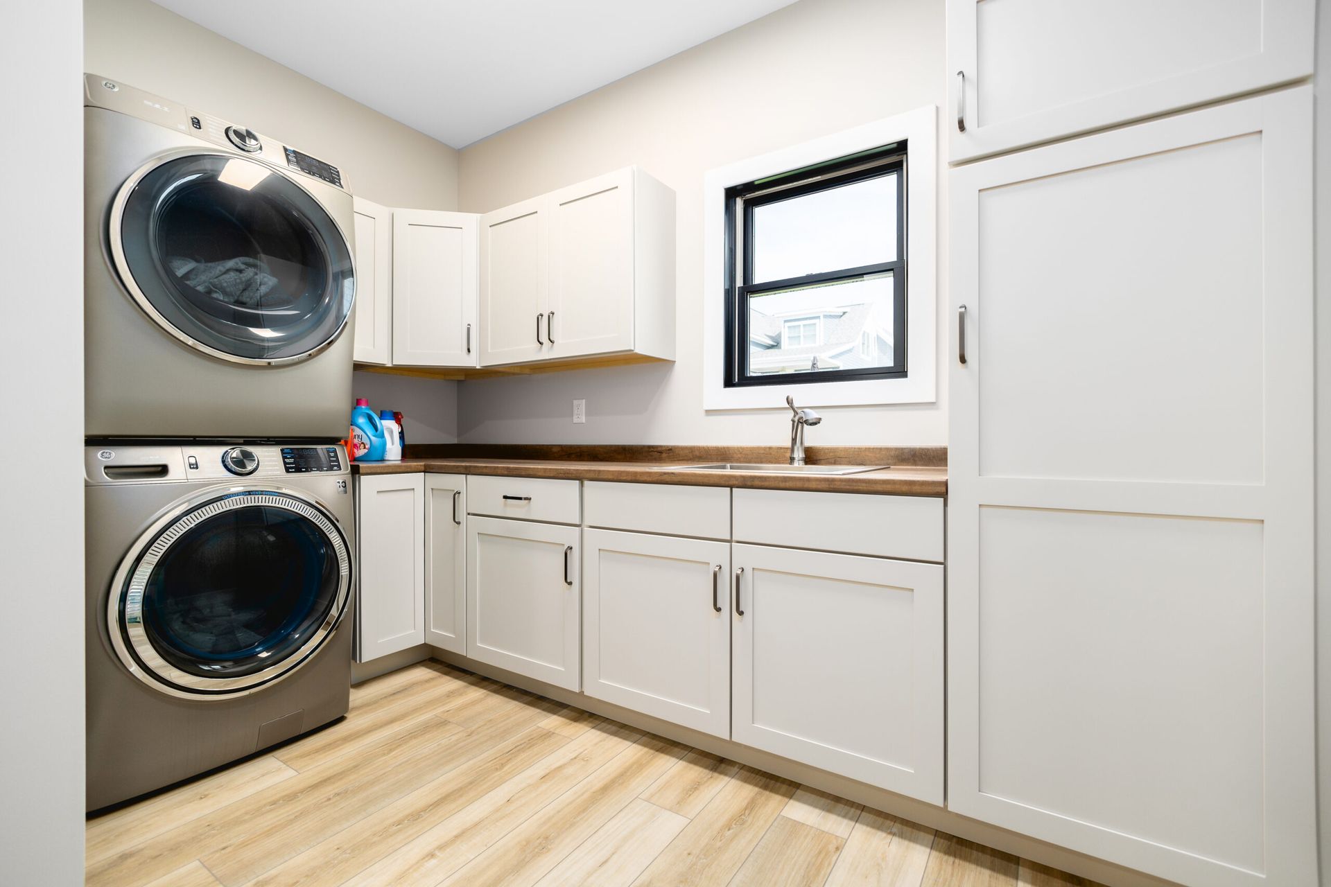 Bright laundry room with stacked washer and dryer, white cabinets, wood floor, and a small window.