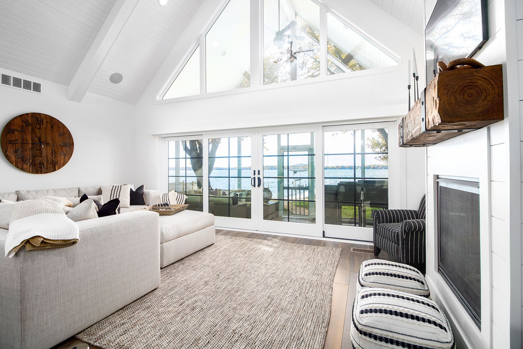 Bright white living room with large windows, gray sofa, patterned rug, and fireplace near a lake view