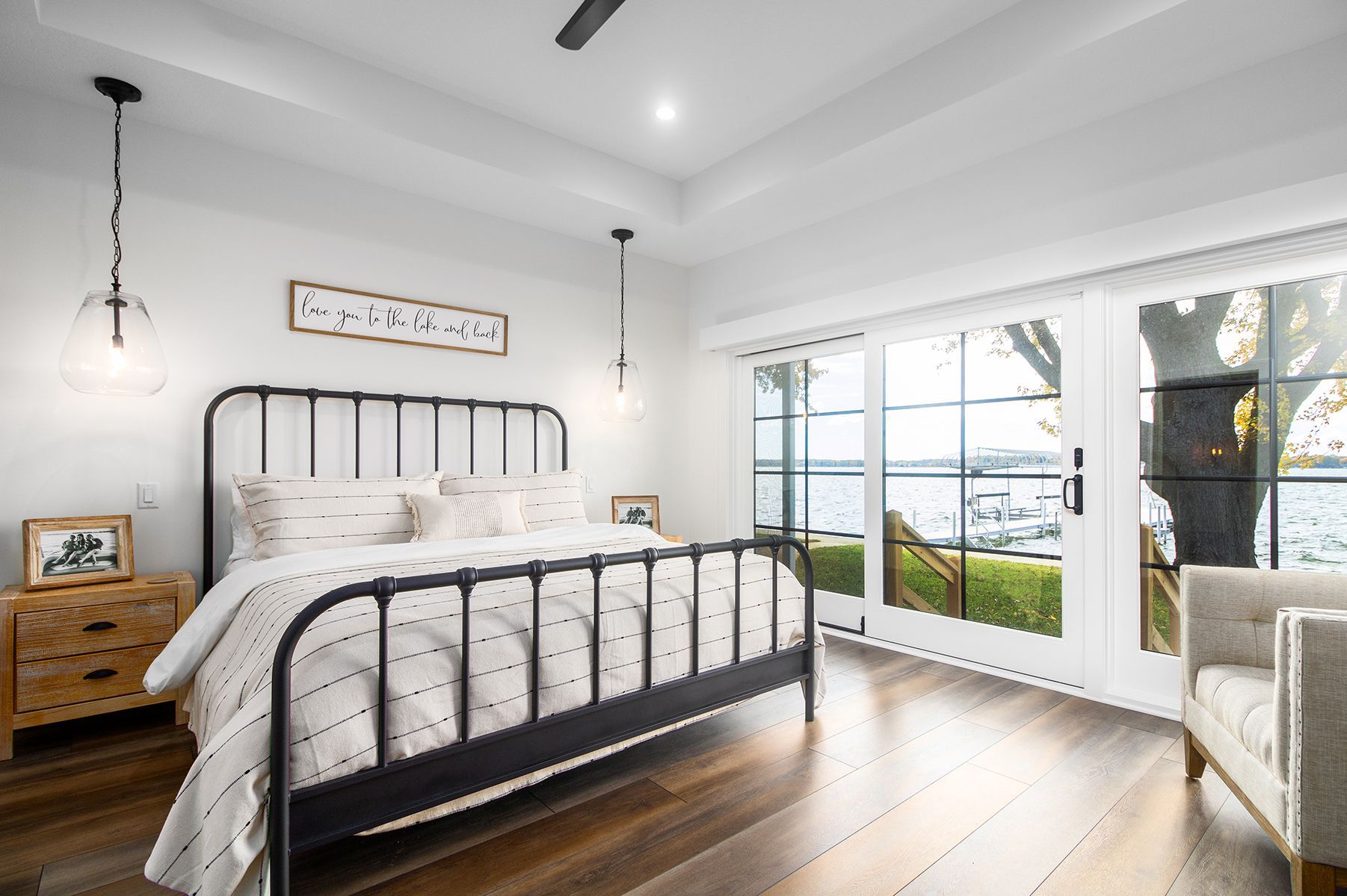 Bright bedroom with black metal bed, white bedding, wooden nightstand, and large windows overlooking a lake.