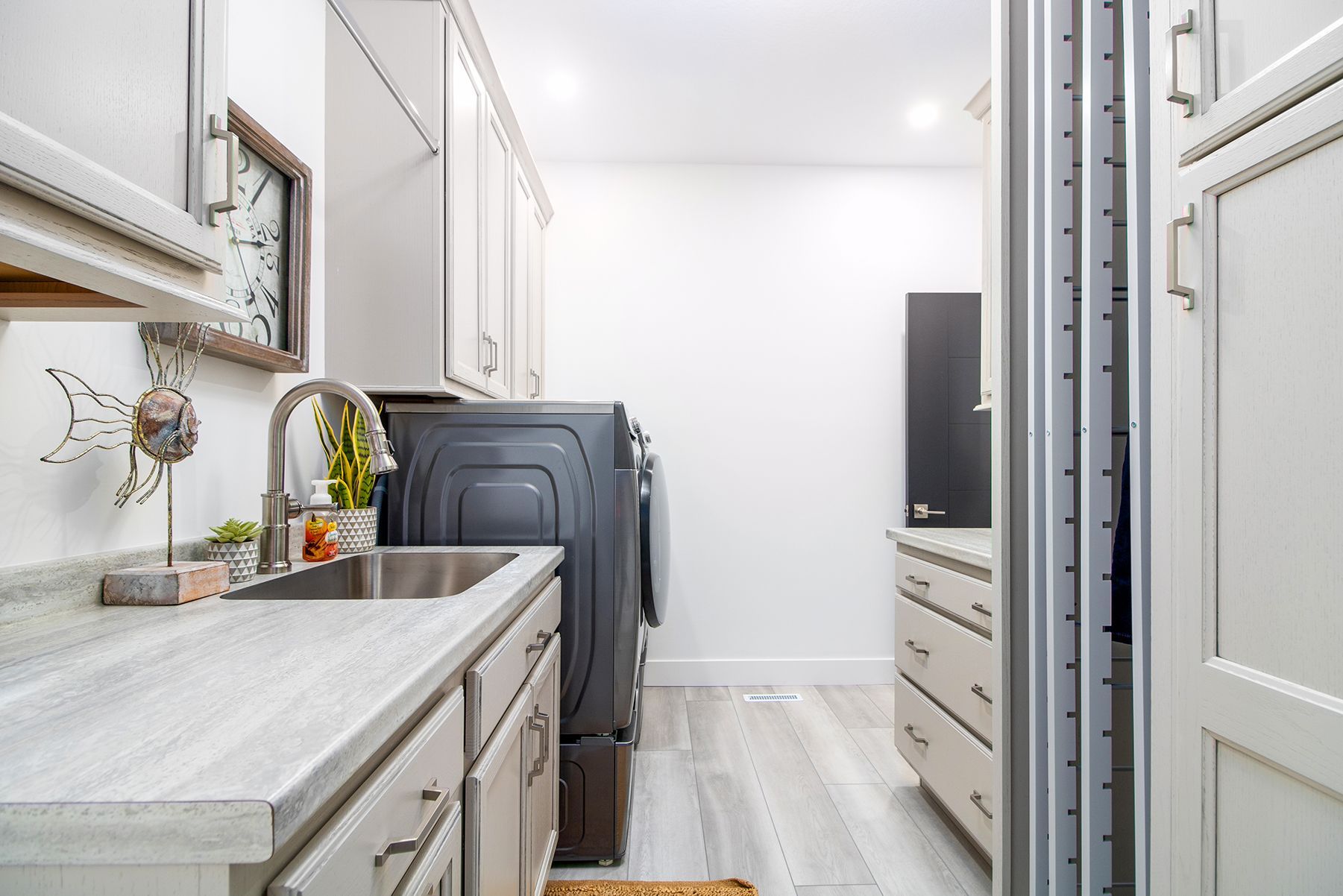 Narrow laundry room with white cabinets, gray countertops, stacked washer and dryer, and a TV at the far end.