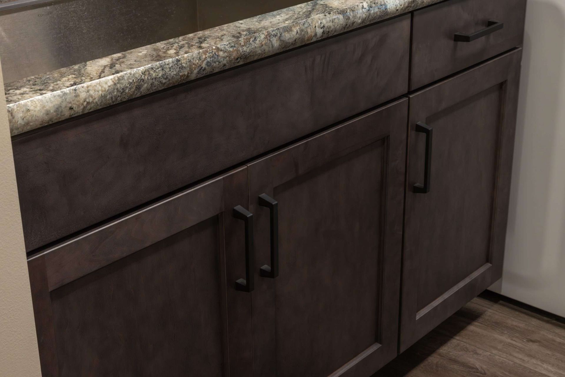 Dark brown kitchen cabinet with black handles and speckled countertop, next to a white appliance.