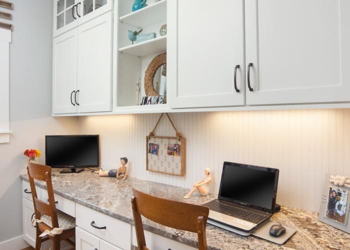 Built-in home office desk with two laptops, white cabinets, and granite countertop under wall shelves