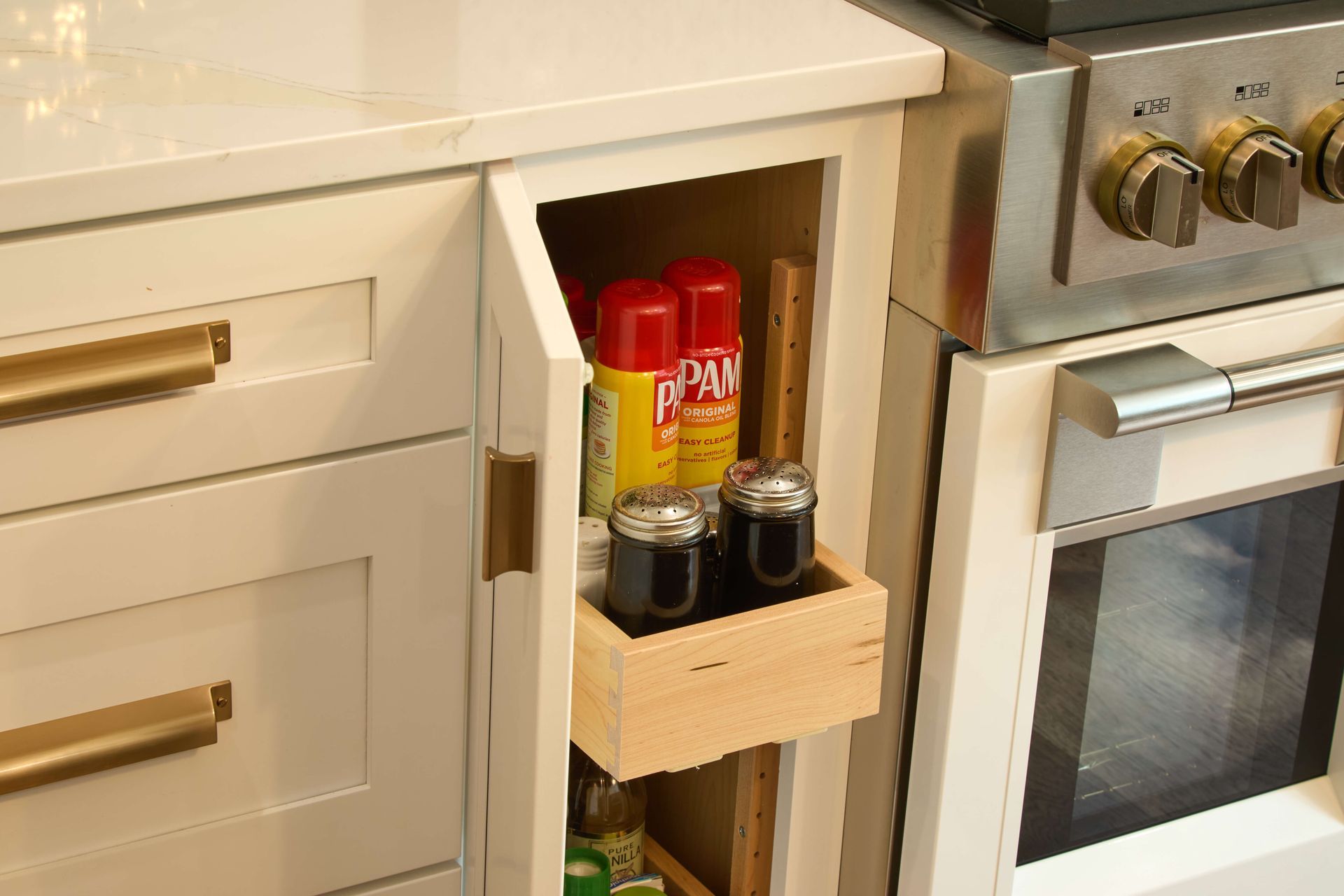 Pull-out kitchen spice rack beside an oven, holding jars and cans in a white cabinet.