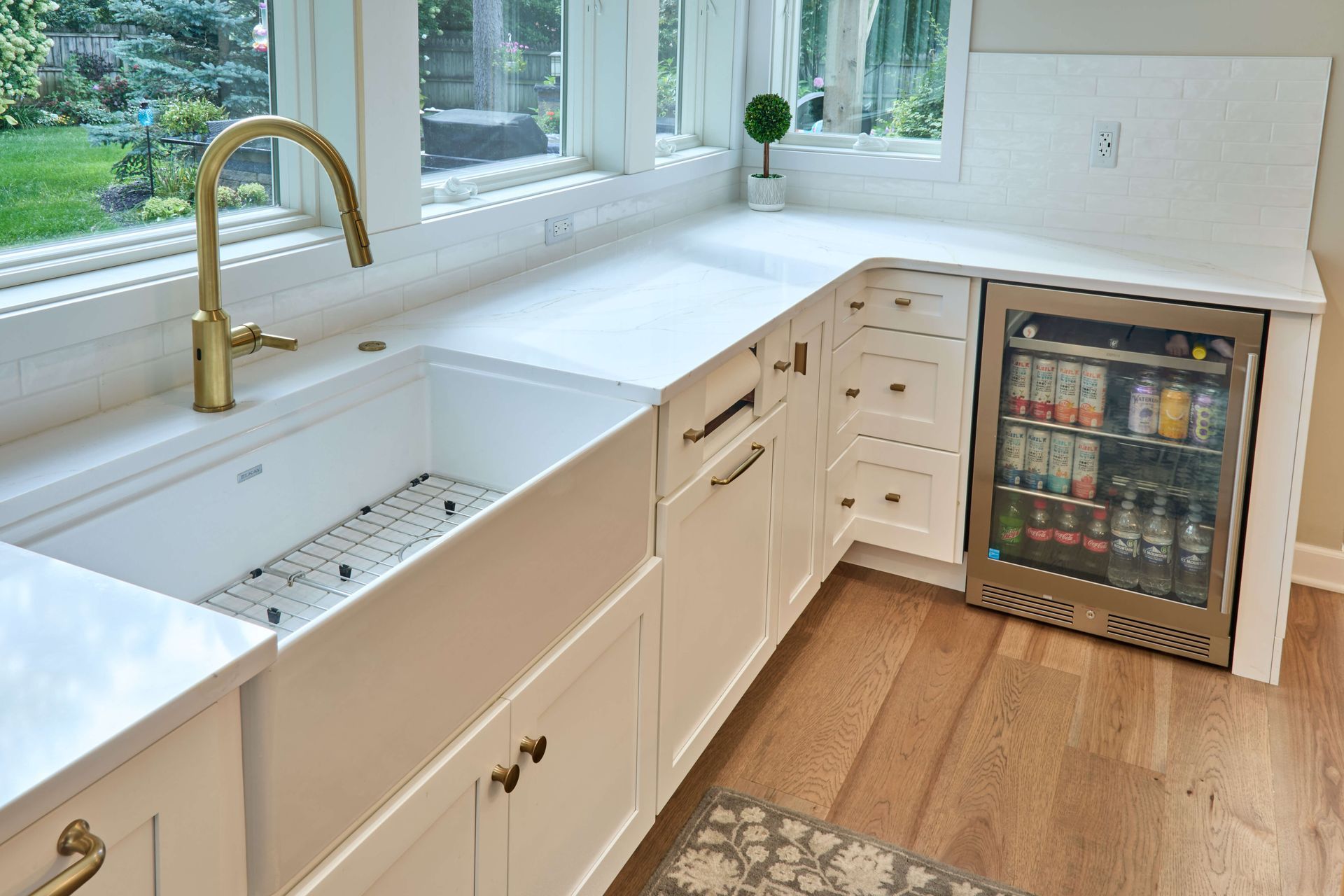 Bright kitchen with white cabinets, farmhouse sink, gold faucet, and under-counter beverage fridge beside a window.