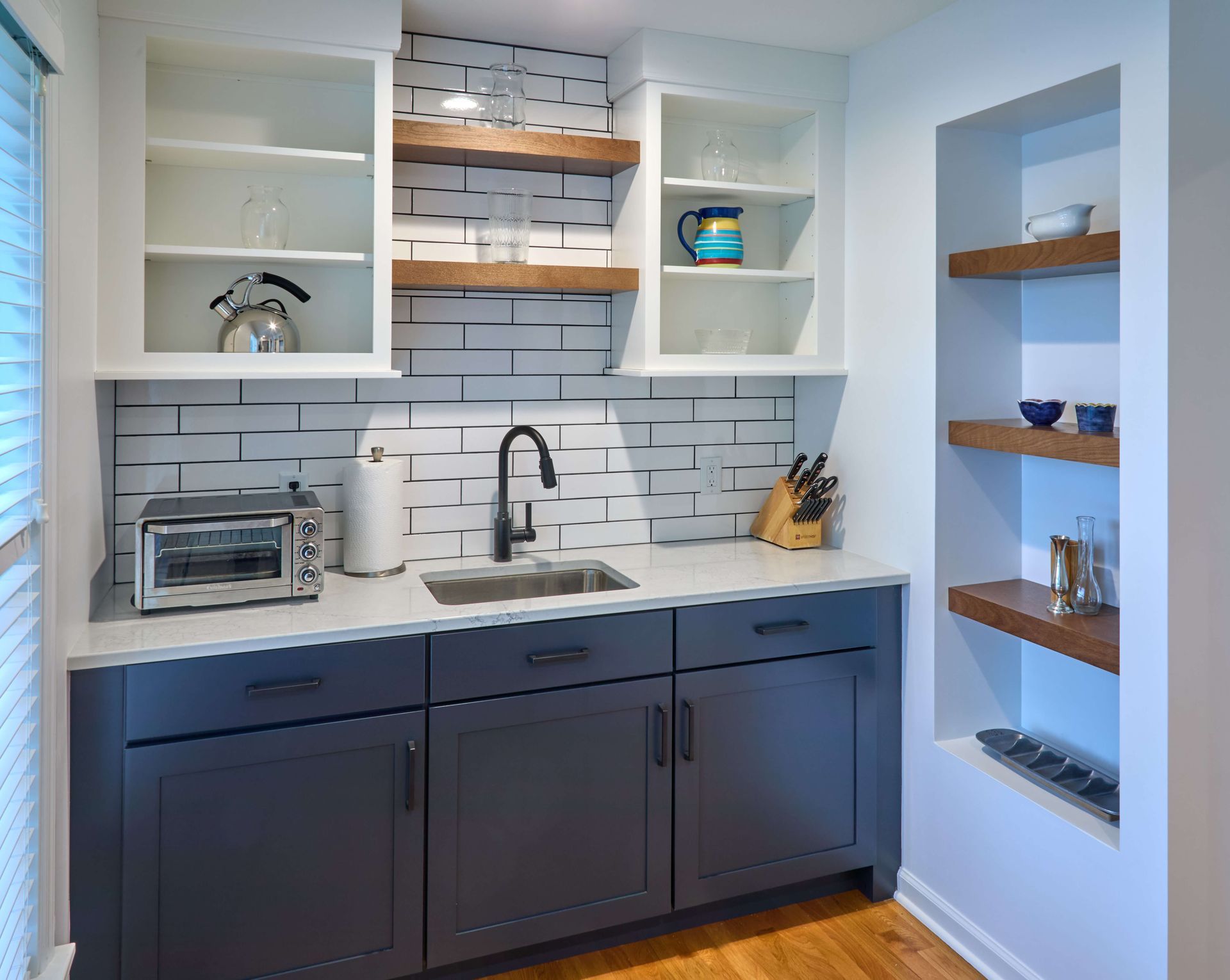 Modern kitchenette with navy cabinets, white subway tile, open shelves, and a stainless toaster oven.