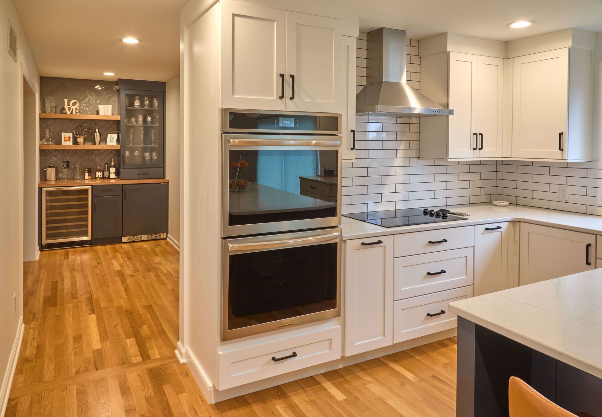 Modern kitchen with white cabinets, stainless appliances, wood floors, and a bar area in the background