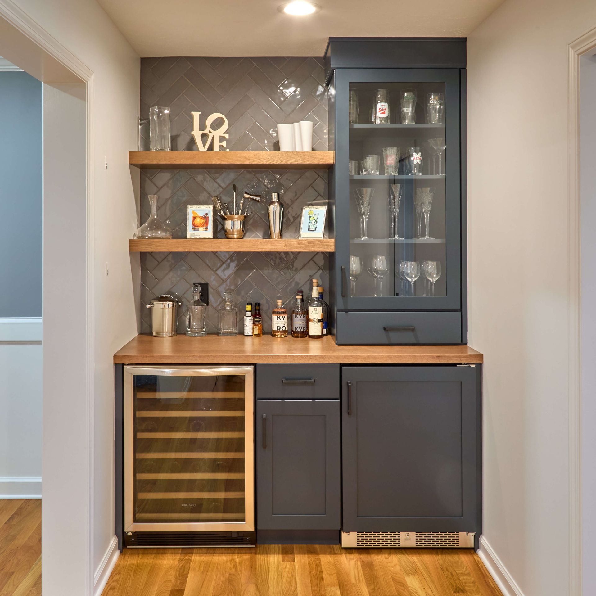 Built-in bar cabinet with dark wood shelves, wine fridge, glassware, and bottles in a home alcove