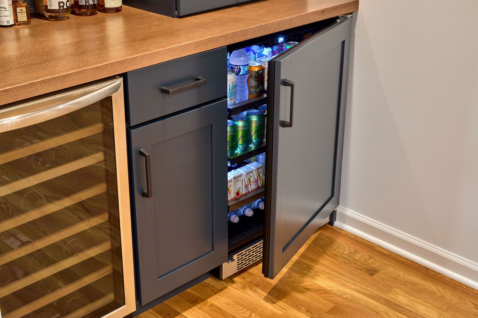 Small under-counter beverage fridge with an open black door in a kitchen, showing colorful cans inside.