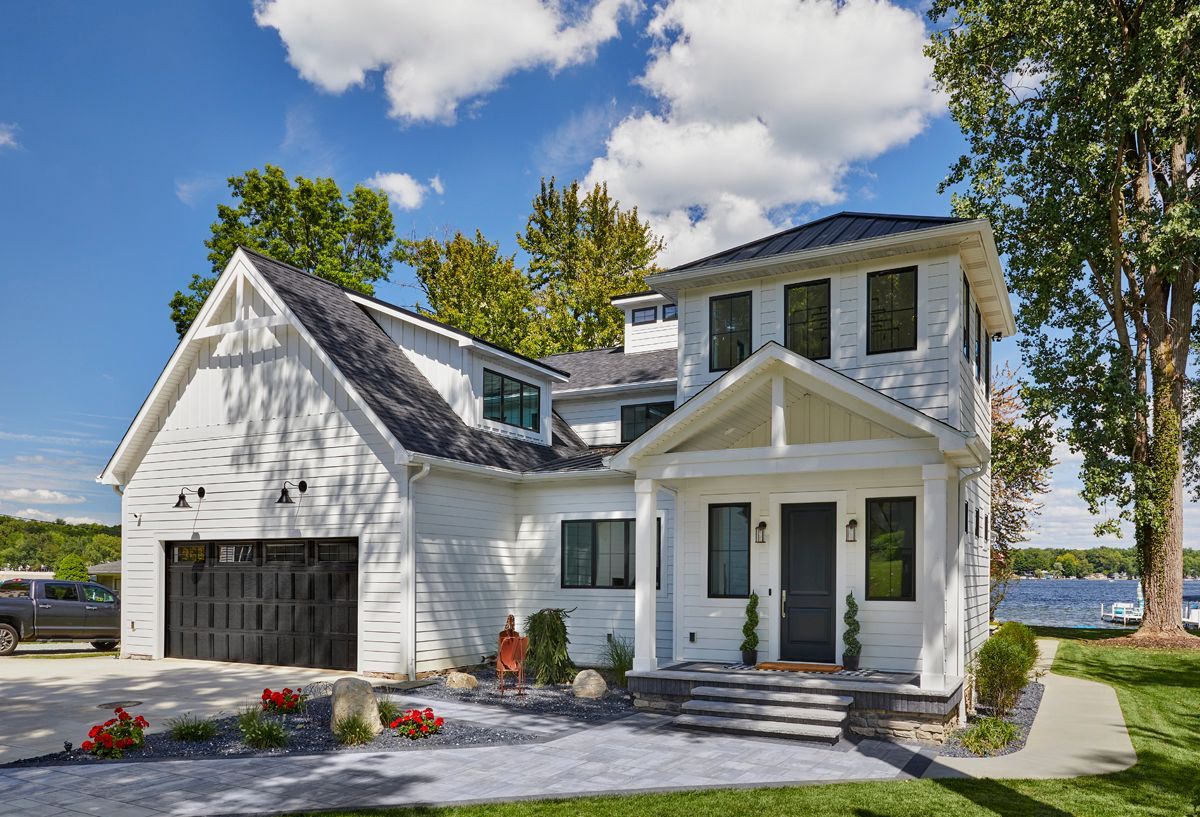 White two-story house with black garage door, front porch, and stone driveway beside water under a sunny sky