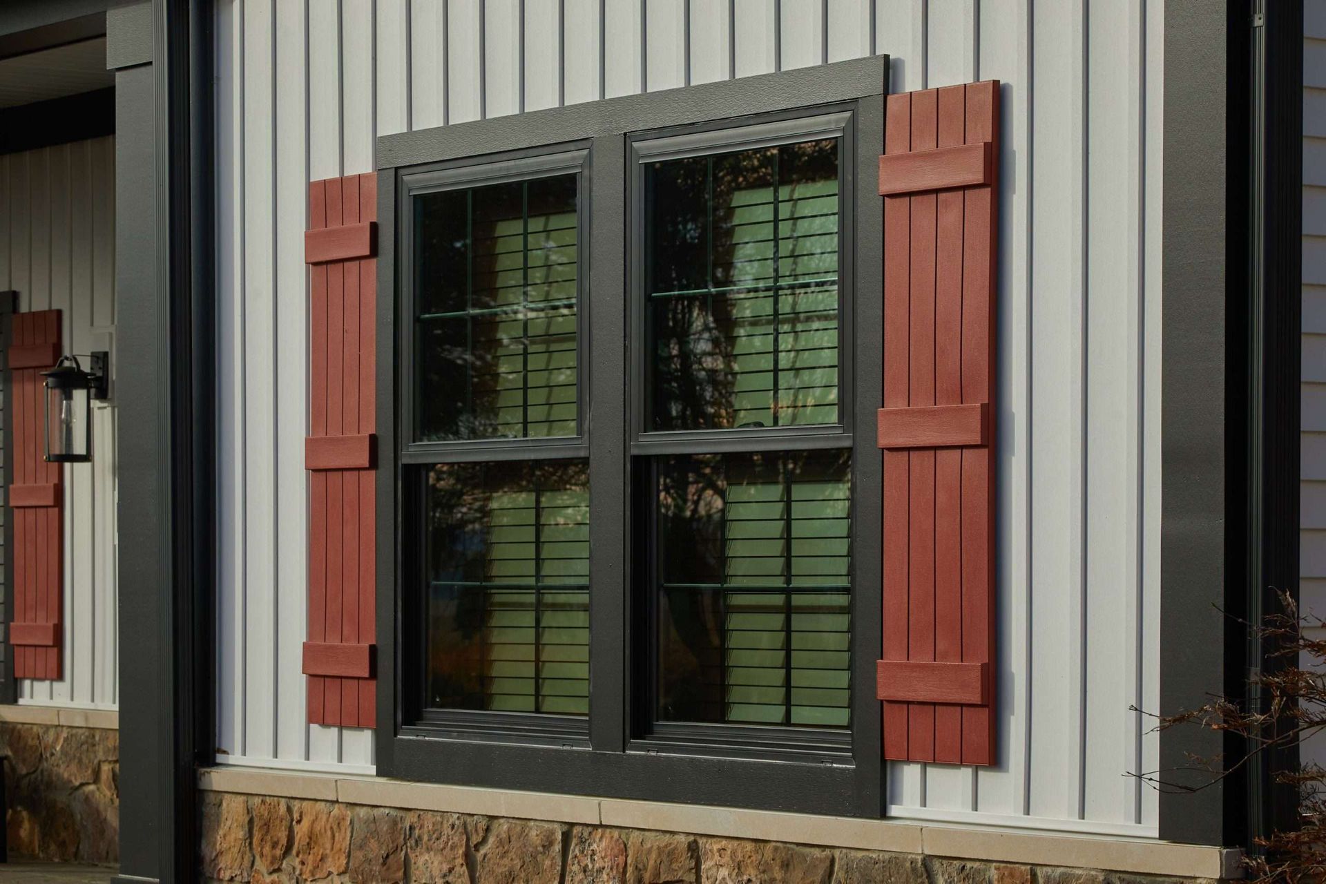 Black-framed window with maroon shutters on gray siding and stone base.
